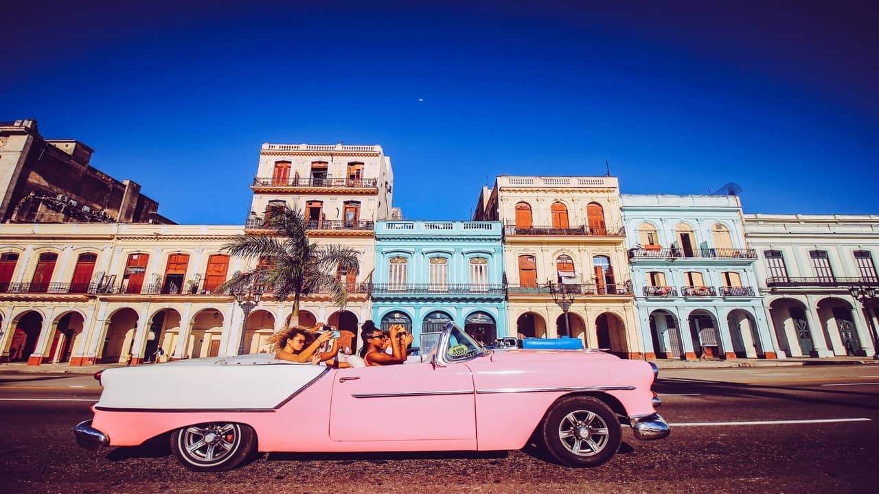 Classic pink convertible driving past colorful colonial buildings in a vibrant street.
