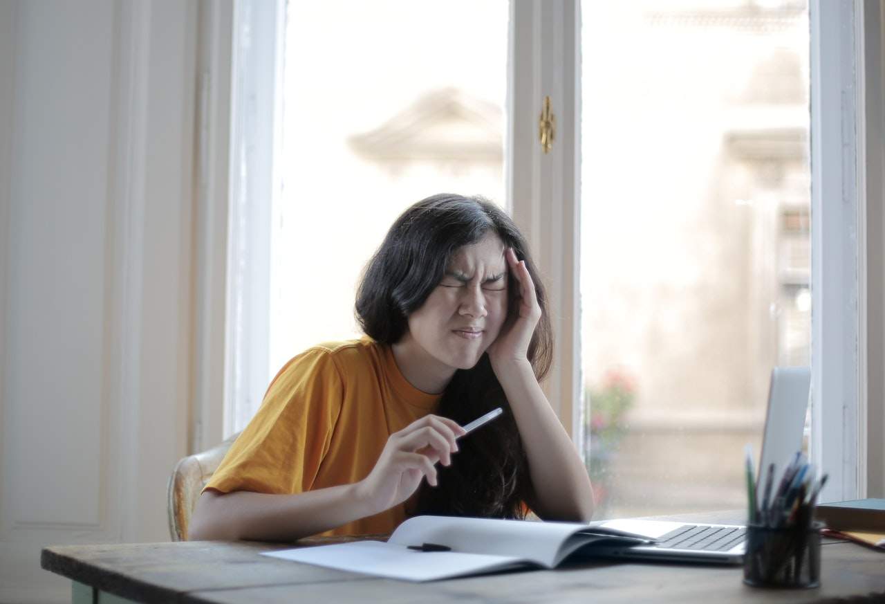 A young woman, appearing frustrated, holds her head while sitting at a desk