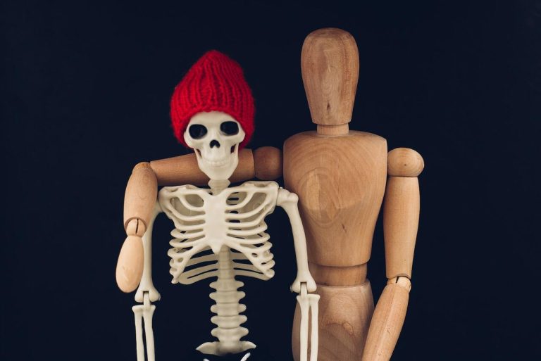 A wooden mannequin with its arm around a small skeleton wearing a red knitted hat