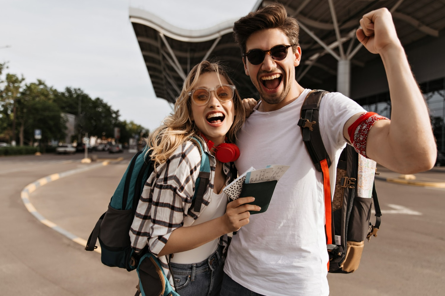 A happy couple celebrating with backpacks and passports outside an airport.