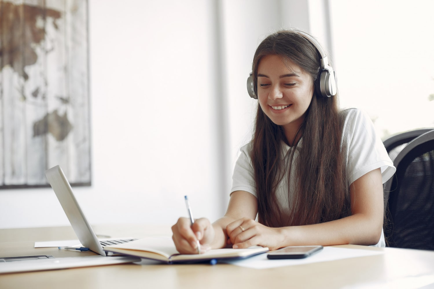 A young woman wearing headphones is sitting at a desk, smiling while writing in a notebook.