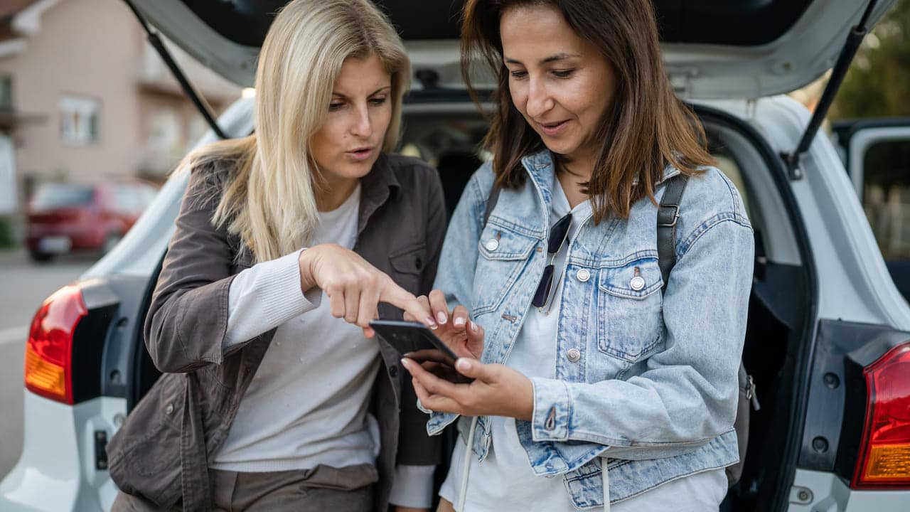 Two women looking at a smartphone near a parked car