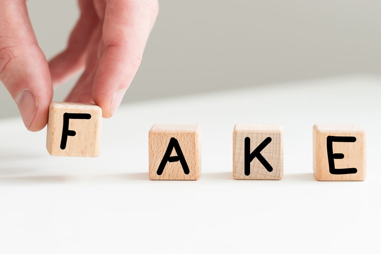 Wooden blocks spell "FAKE," with a hand adjusting the "F."