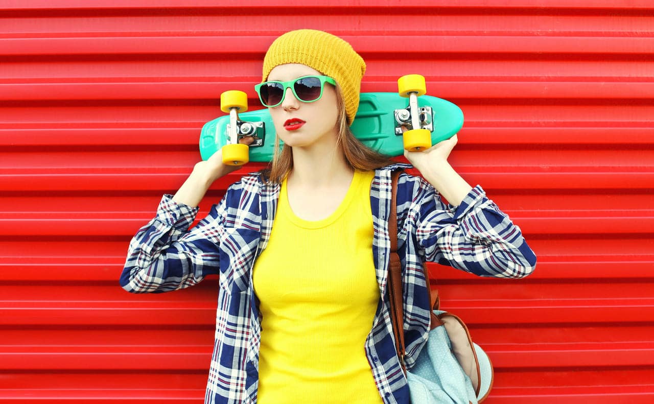 Young woman in sunglasses holding a skateboard against a red background.