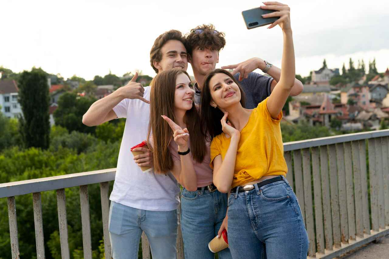 Group of four friends taking a selfie outdoors, smiling and making hand gestures