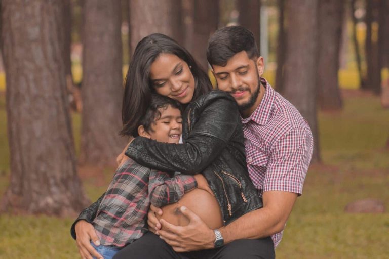 Family outdoors hugging, father holding mother’s pregnant belly.