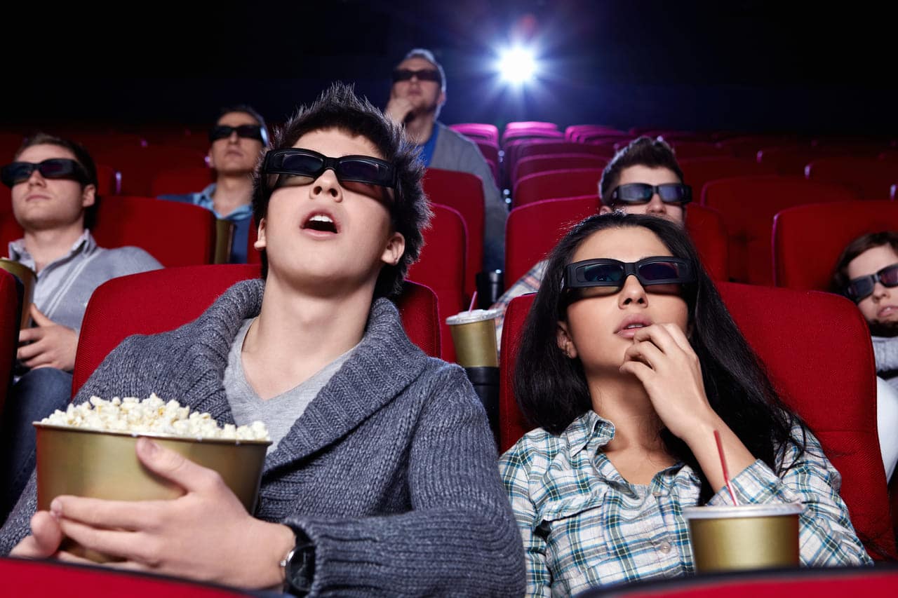 People in a theater wearing 3D glasses, eating popcorn.