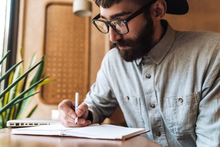 Man with glasses writing in a notebook at a desk