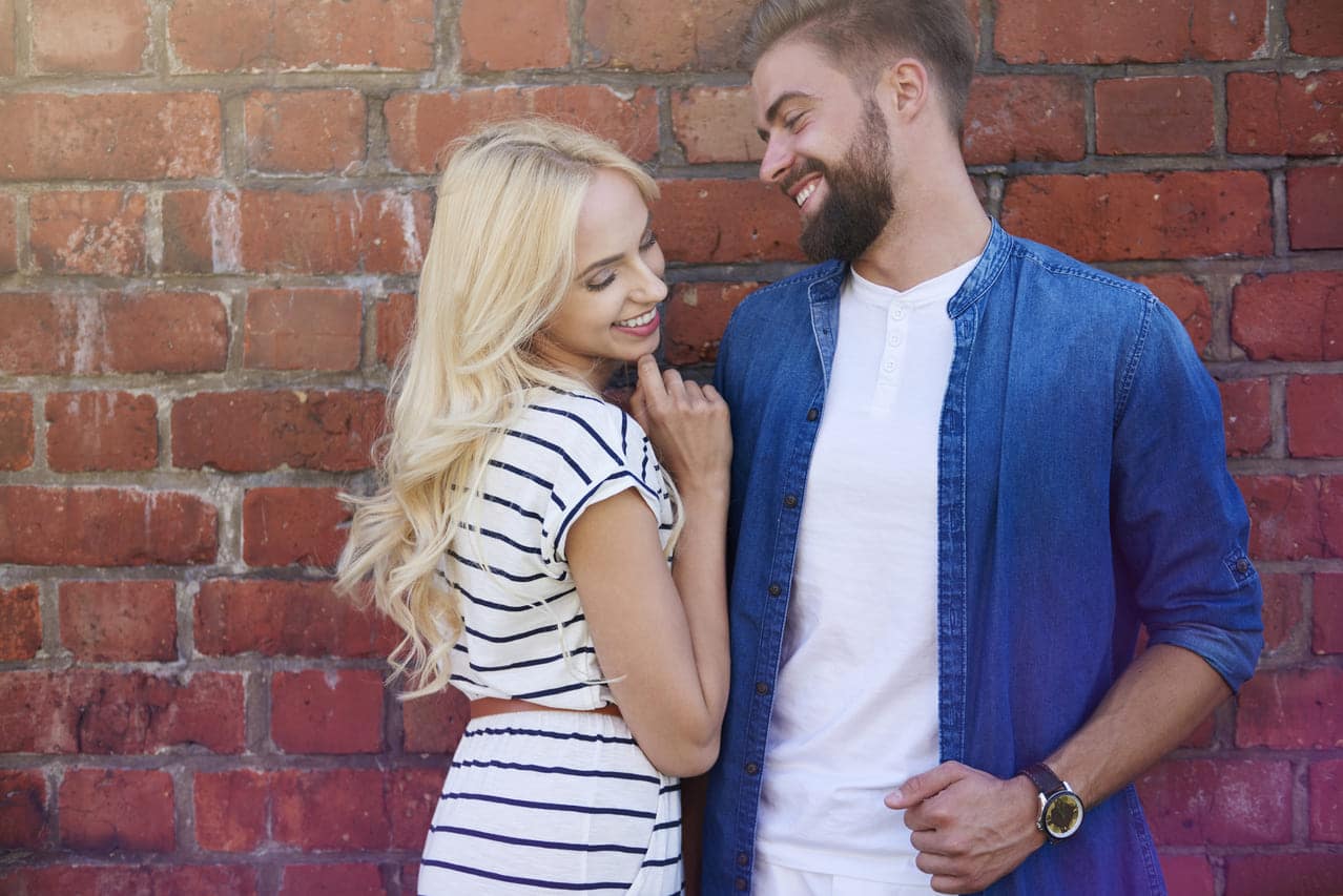 Smiling couple leaning against a brick wall, sharing a tender moment