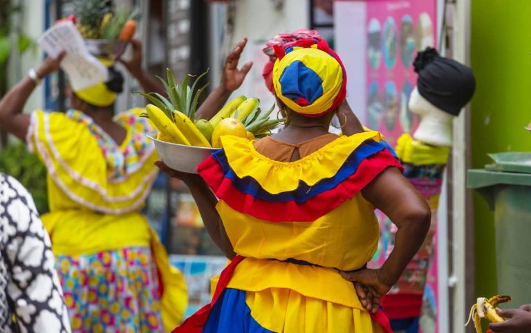 A woman in traditional vibrant clothing carries a bowl of fruits