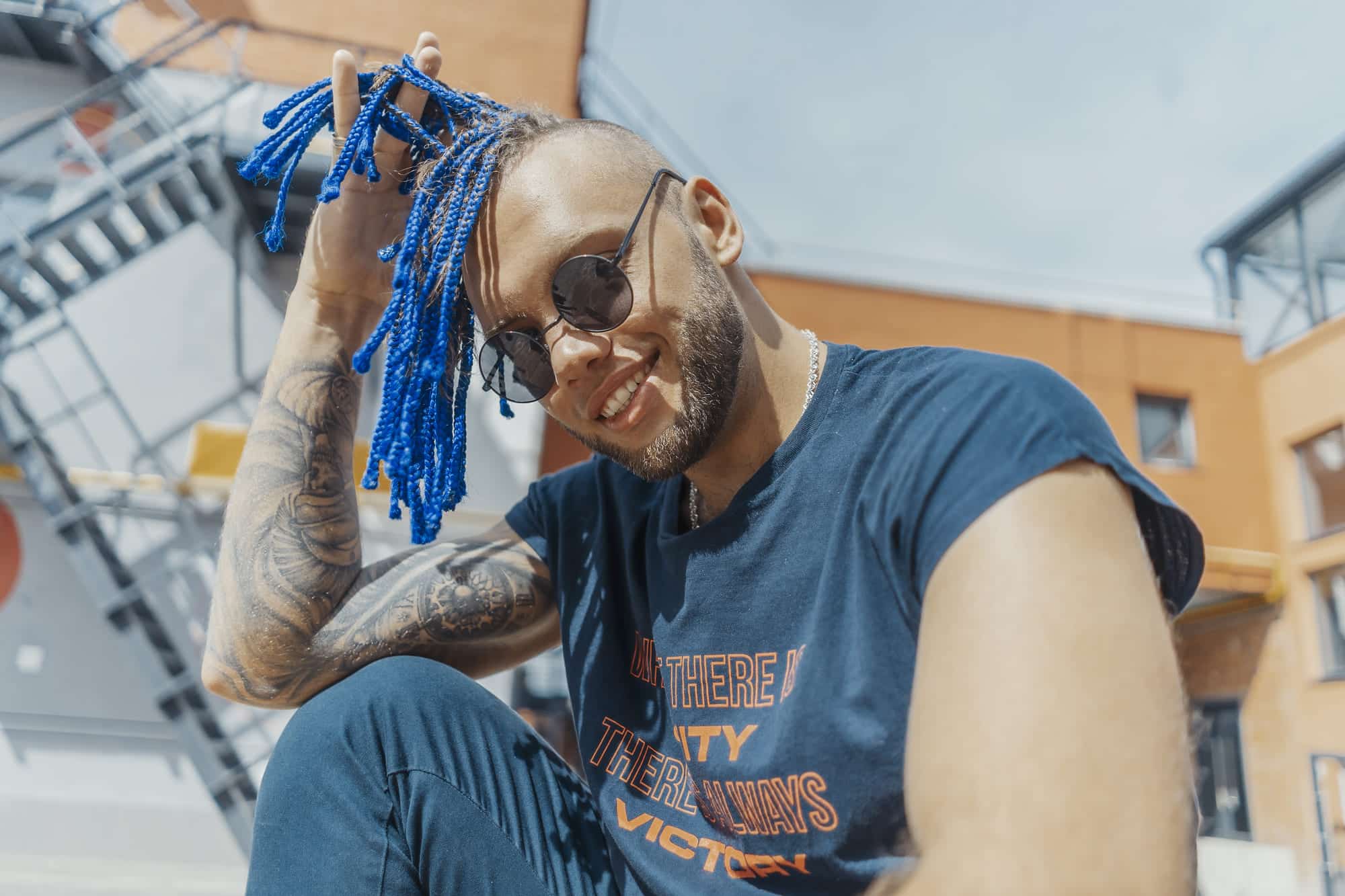 A man with sunglasses and blue braids, smiling outdoors with a tattooed arm.