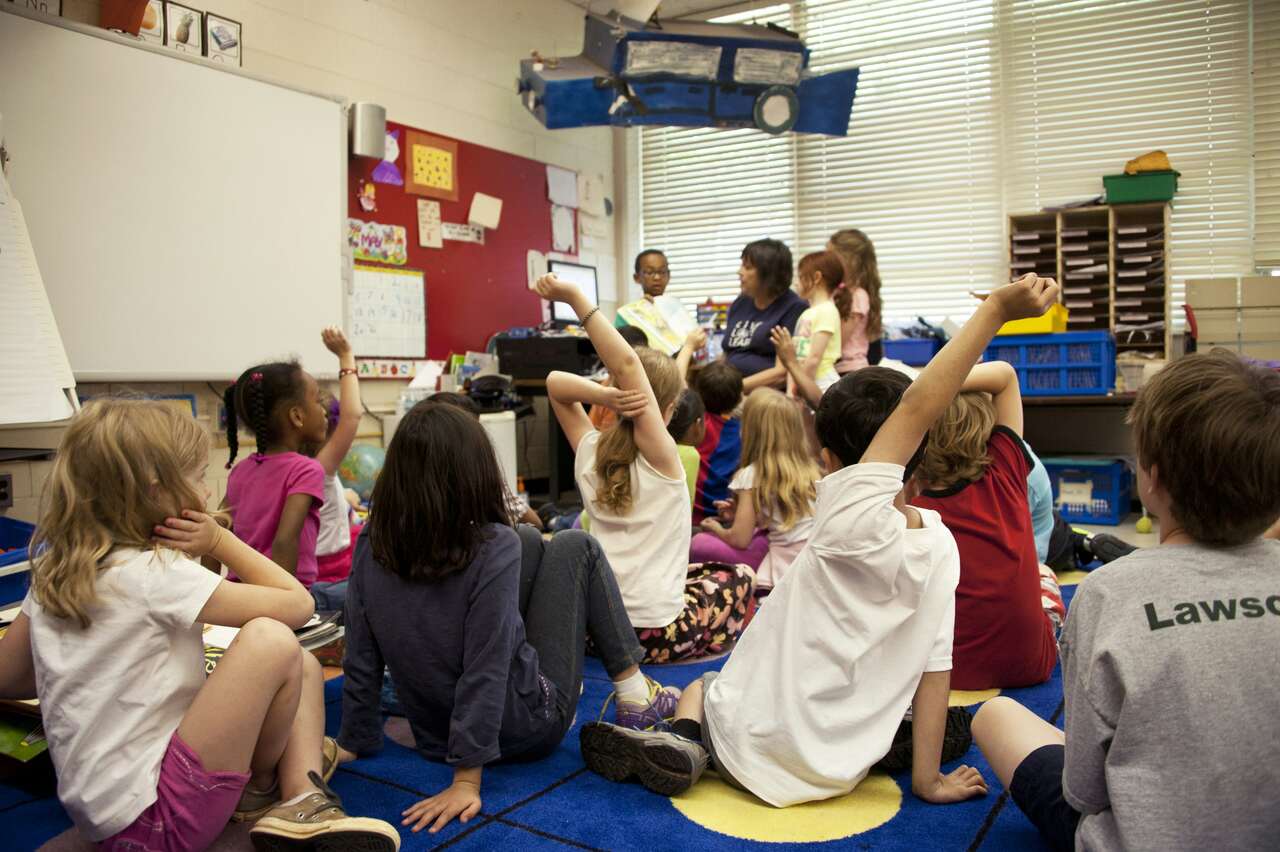 Children sitting on the classroom floor, raising hands to participate in a lesson led by a teacher.