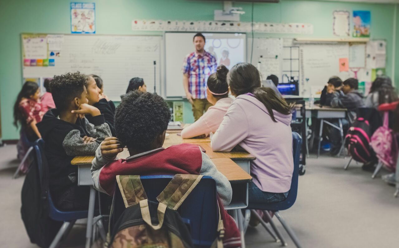 Middle school students seated at desks, attentively listening to a teacher presenting in front of the classroom