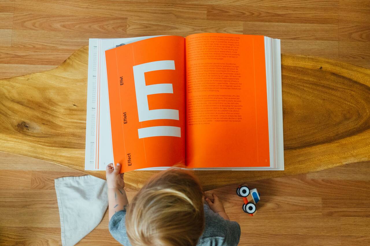 Young child standing at a table, looking at an open book with a bright orange page and bold design.