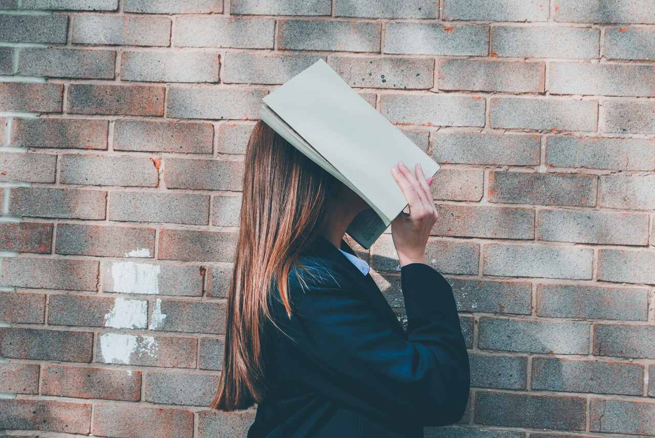 A woman standing against a brick wall with a book covering her face, holding it up as if in frustration or fatigue.