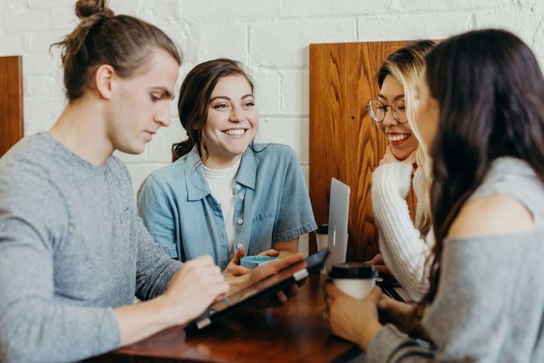 A group of four young adults sitting together at a table, smiling and discussing something, with a tablet and a laptop on the table, in a casual and friendly environment.