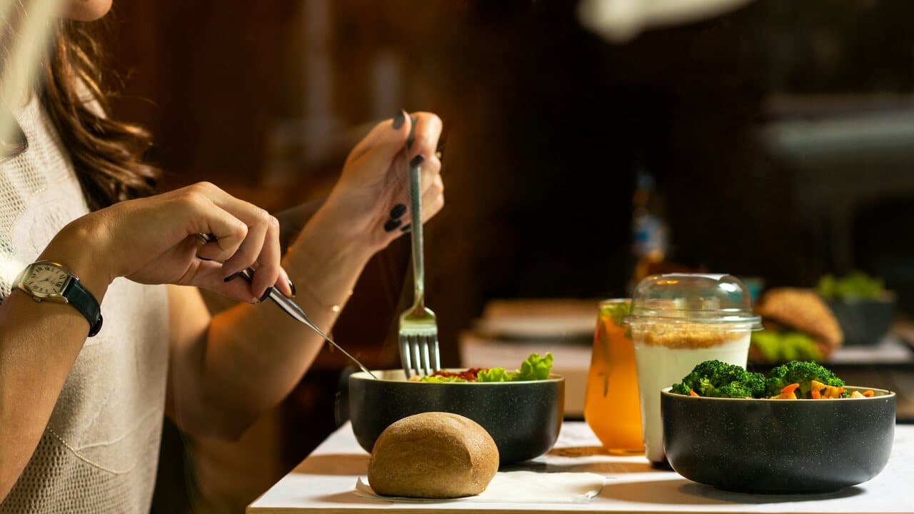 Person eating a meal at a table with a bowl of salad, a small loaf of bread, a glass of juice, and a yogurt parfait, using a fork and knife.