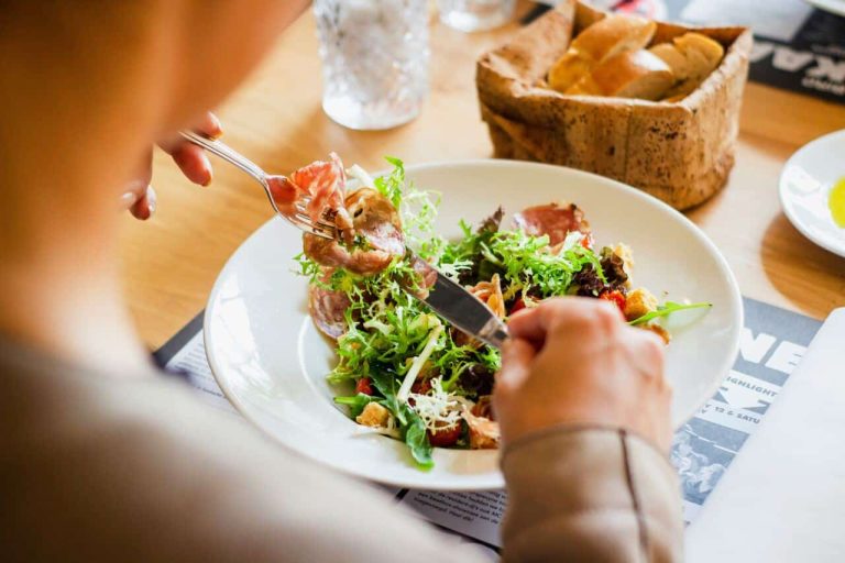 Person eating a fresh salad with a fork and knife, with a basket of bread and a drink in the background on a wooden table.