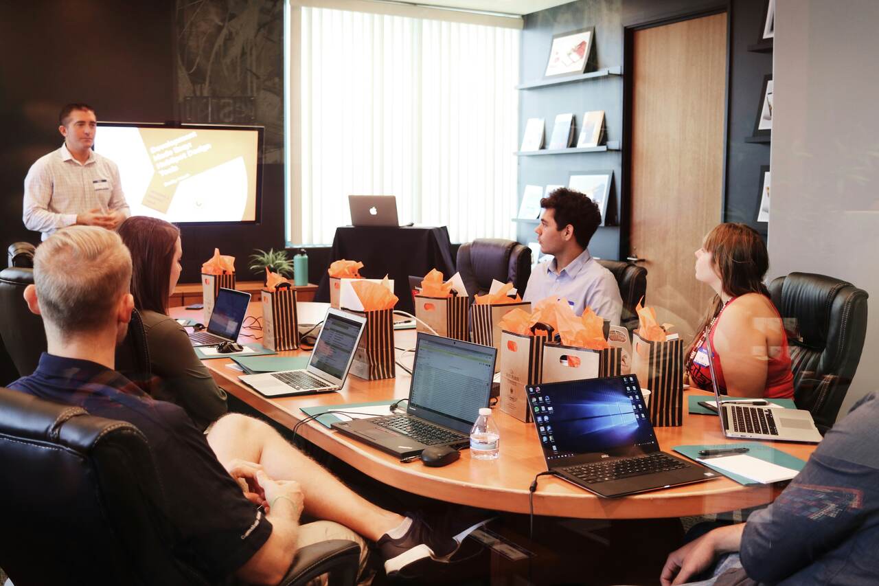 A group of people seated around a conference table with laptops, listening to a presenter standing by a screen, delivering a presentation in a modern office space.