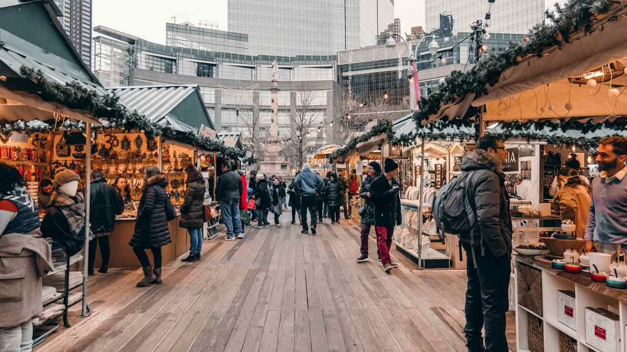 Busy outdoor holiday market with shoppers browsing through stalls decorated with greenery and lights, selling handmade crafts, ornaments, and gifts, set against a backdrop of tall buildings.