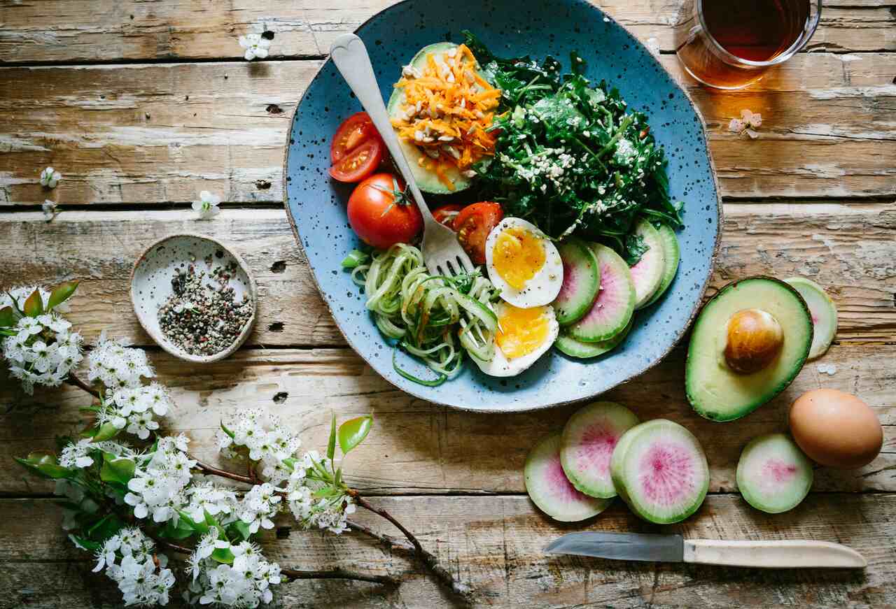 Top view of a healthy meal on a blue plate, including boiled eggs, tomatoes, zucchini noodles, avocado, and leafy greens, placed on a rustic wooden table alongside sliced radishes, avocado, and a bowl of seeds.
