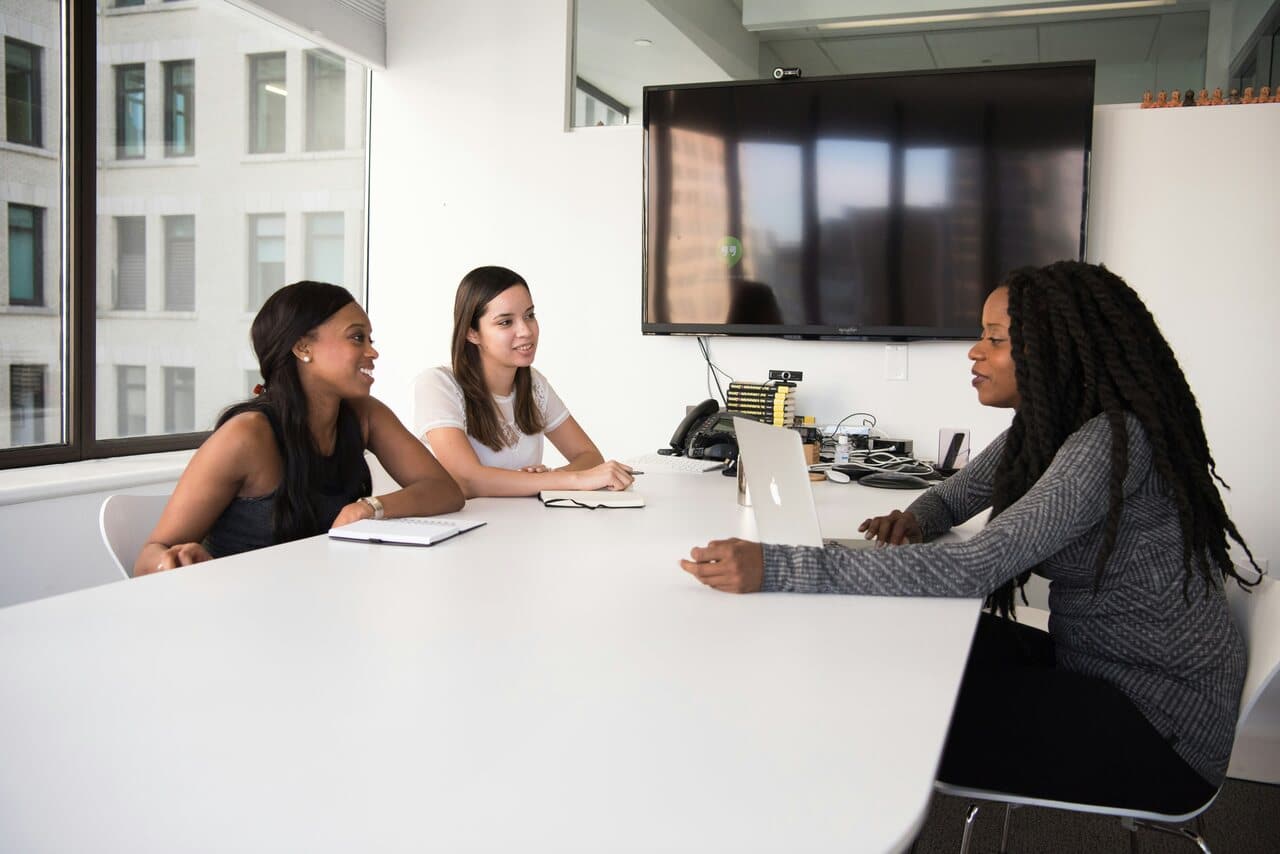 Three women sitting at a table in a bright office space, engaged in a discussion with a laptop in front of them, and a large screen on the wall behind.